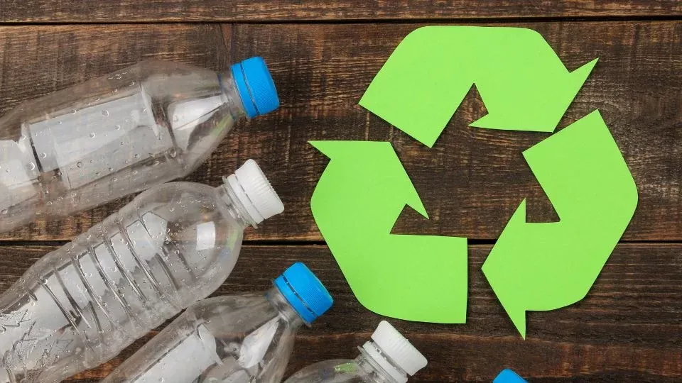 Clear plastic water bottles with blue and white caps arranged next to a large green paper recycling symbol on a dark wood surface.