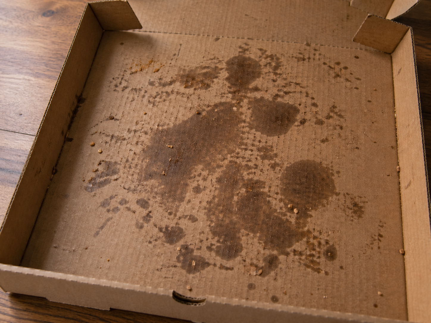 An empty pizza box opened on a wooden surface, the cardboard interior heavily stained with dark grease blotches and scattered food crumbs.