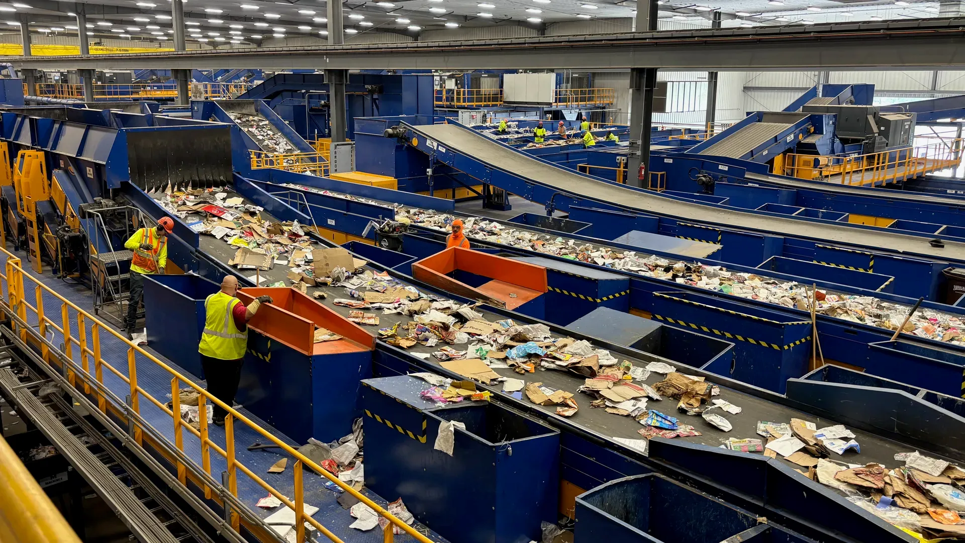 Workers in high-visibility vests sort recyclables on a network of blue conveyor belts in a large industrial recycling facility.